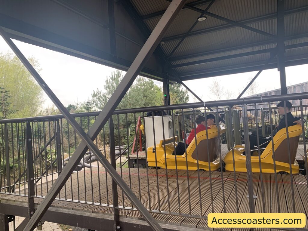 Roller coaster station platform with a yellow roller coaster train. The train is stationary under a metal roof, and there are three people seated in the front two rows, waiting for the ride to begin. The platform is surrounded by metal railings
