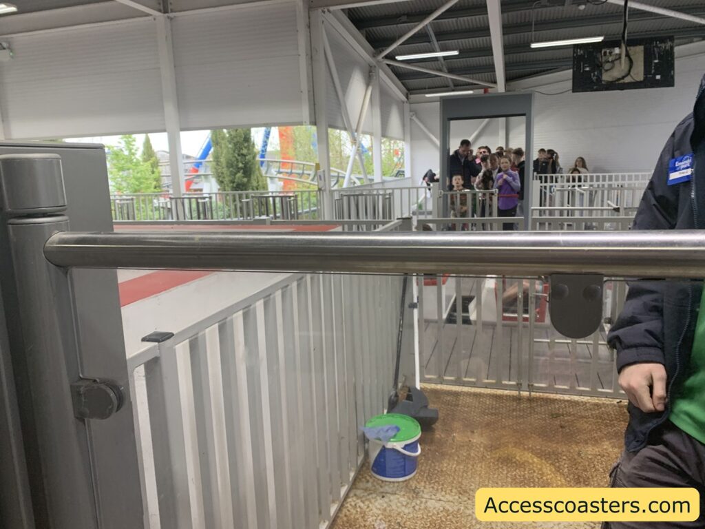 Amusement park ride queue area with metal railings and a turnstile in the foreground. There is a group of people, including children, waiting behind a gate in the background. Part of a staff member wearing a dark jacket and green shirt is visible on the right side of the image. The floor is metallic and slightly dirty, with a mop and a bucket placed near the railing. Outside, some trees and parts of a roller coaster track can be seen. 