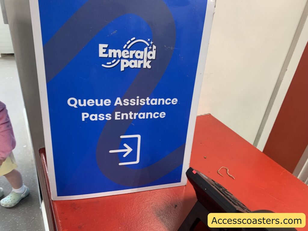 A blue sign with white text that reads "Emerald Park Queue Assistance Pass Entrance" and an arrow pointing to the right. The sign is placed on a red counter. In the bottom right corner,