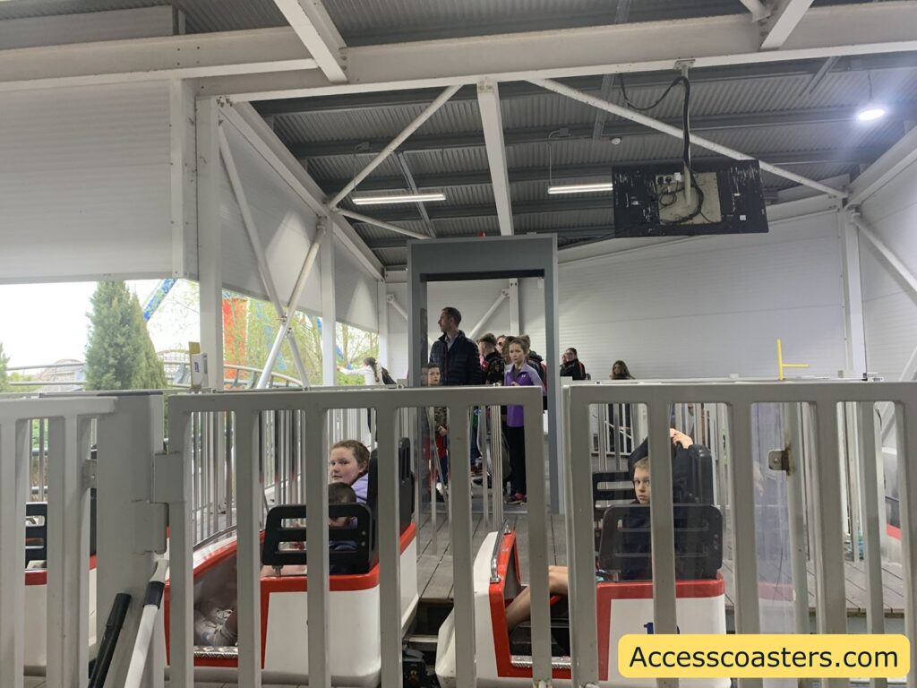 Roller coaster loading station with children seated in small white and red coaster cars, waiting for the ride to begin. There are metal safety gates in front of each car, and a group of people, including adults and children, are standing in line behind the cars.