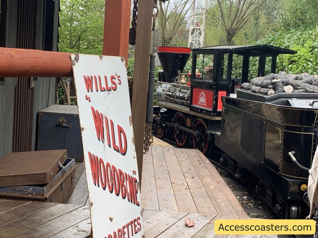 Old-fashioned steam train at a station platform. The train is black with red and gold accents, and its tender is filled with logs, suggesting it is a wood-burning locomotive. Next to the train, there is a large, weathered sign leaning against a post that reads in red letters: "WILLS'S 'WILD WOODBINE' CIGARETTES." The platform is wooden, and there are a couple of old suitcases stacked nearby,