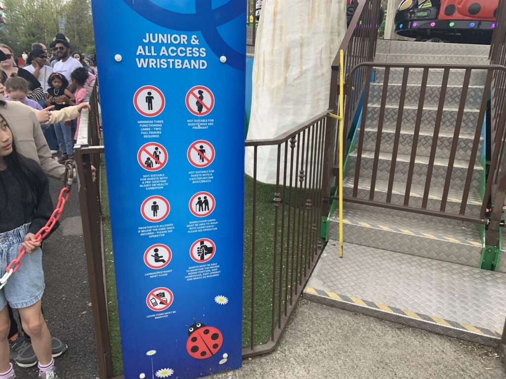 Blue signboard at an amusement park ride entrance. The sign lists safety rules and requirements for the ride, titled "JUNIOR & ALL ACCESS WRISTBAND." There are nine circular icons with red borders and diagonal slashes, each representing a restriction or rule. The rules are:

Minimum three functioning limbs - two full arms required.
Not suitable for guests who are pregnant.
Not suitable for guests with pre-existing health conditions.
Not suitable for suit, limb, arm cast/fracture boot/brace.
Prosthetics allowed if secure and operator has assessed.
Wheelchair users must be able to walk unaided; wheelchair must be left at the ride exit.
Lap bar/restraint must close.
Guests must be able to maintain an upright position and brace as required.
Loose items must be removed.

To the right of the sign is a metal staircase with a brown railing leading up to the ride platform. People, including children and adults, are waiting in line on the left side of the image.
