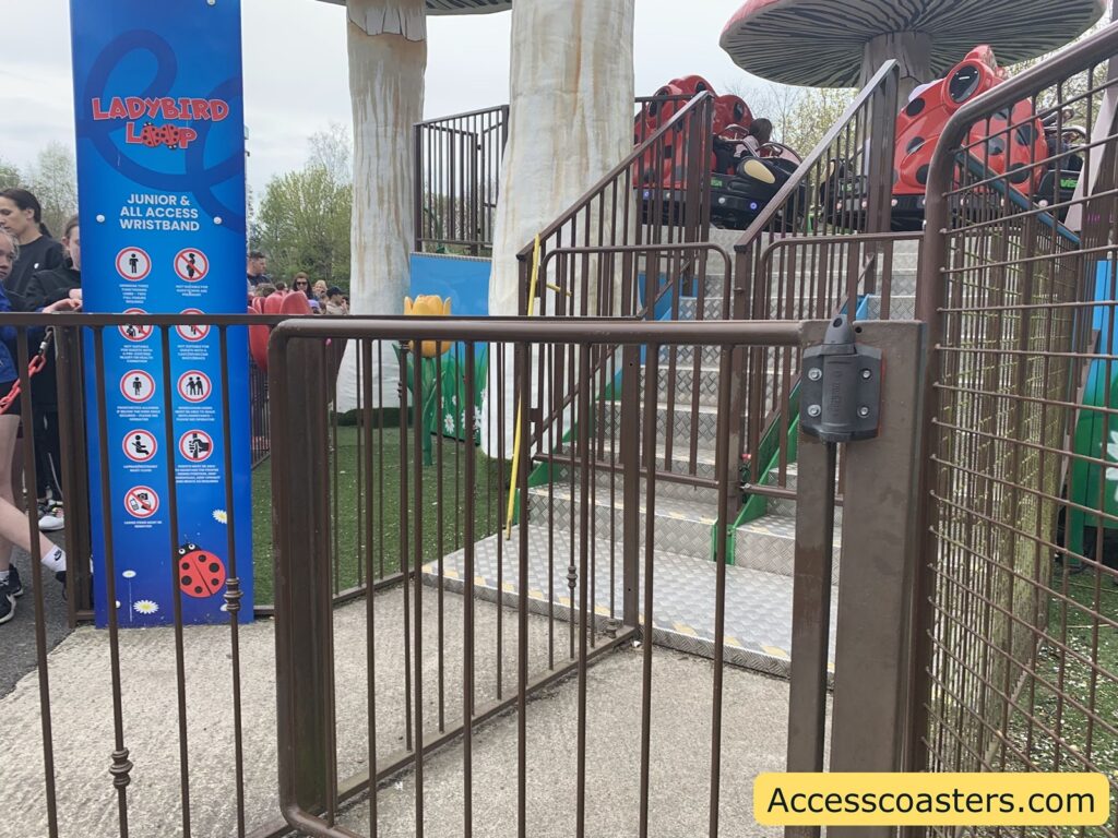 Amusement park ride entrance with a metal gate in the foreground and a blue sign on the left. The sign reads "LADYBIRD LOOP" and lists requirements for a junior and all access wristband, along with several icons indicating rules such as no food or drink, no loose articles, and accessibility information. Behind the gate, there are steps leading up to the ride platform, where ladybird-shaped ride cars are visible. The ride is themed with large mushroom decorations and artificial grass.