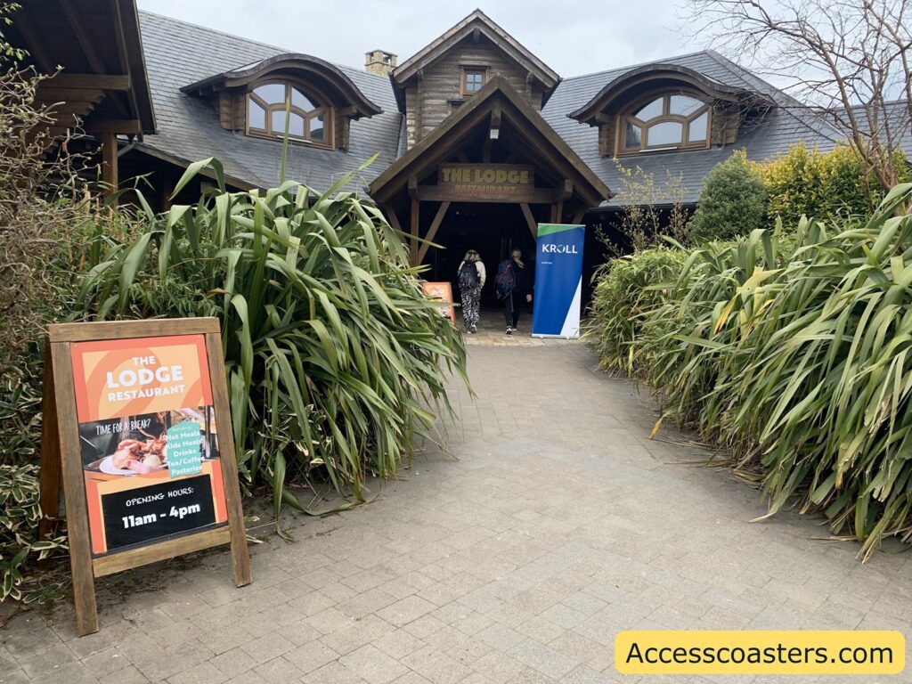 Wooden lodge-style restaurant building with a steep slate roof, surrounded by lush green plants. There is a wide paved path leading up to the entrance, where two people are walking inside. On the left side of the path, there is a wooden-framed sign advertising "The Lodge Restaurant" with opening hours from 11am to 4pm, and mentioning hot meals, kids meals, drinks, coffee, and pastries. Near the entrance