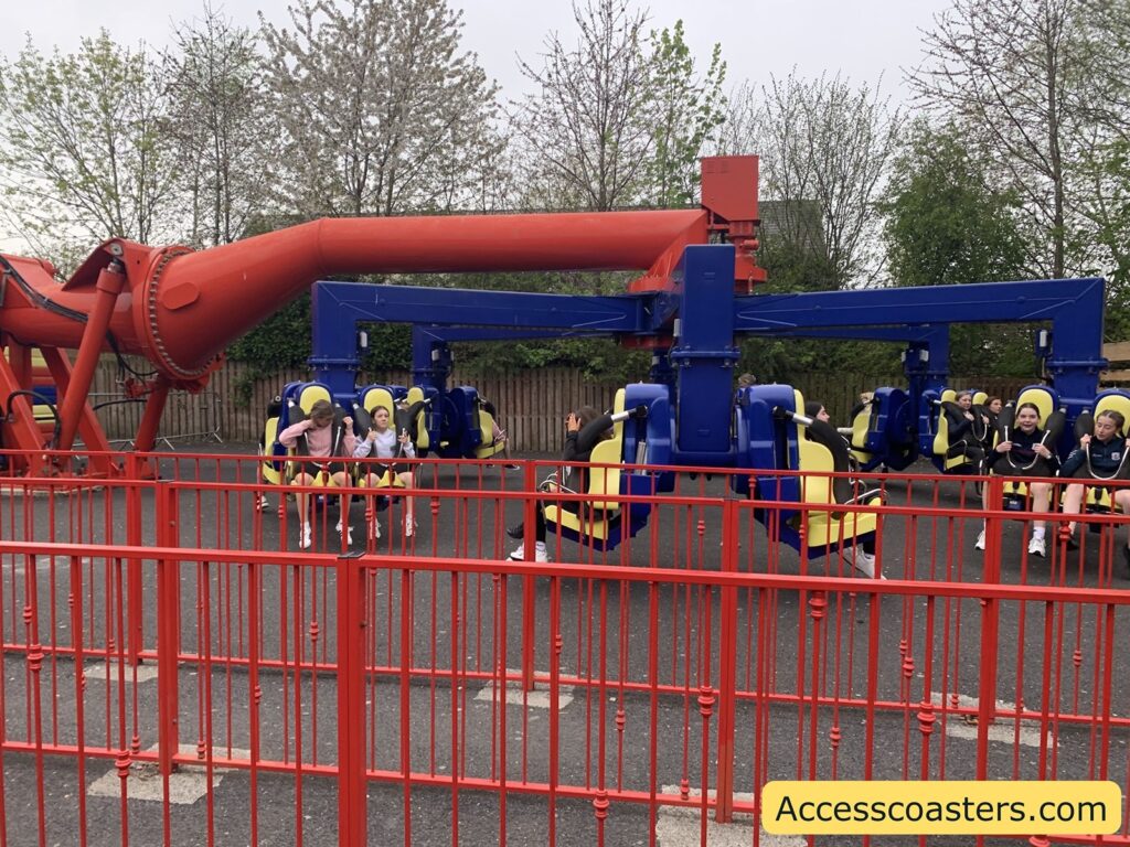 Power surge  ride with several people seated in yellow and blue seats, secured with over-the-shoulder restraints. The ride structure is mostly blue with a large red arm overhead, and it is surrounded by a red metal fence. The people on the ride appear to be waiting for it to start, with some smiling and others looking excited or nervous.