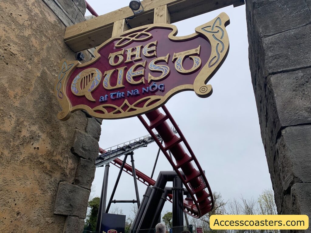 Wooden and metal theme park entrance sign for "The Quest at Tír na nÓg." The sign is ornate, with gold and purple Celtic-style designs, and is mounted between two stone walls. Below the sign, a red roller coaster track curves through the scene, supported by black and silver beams.