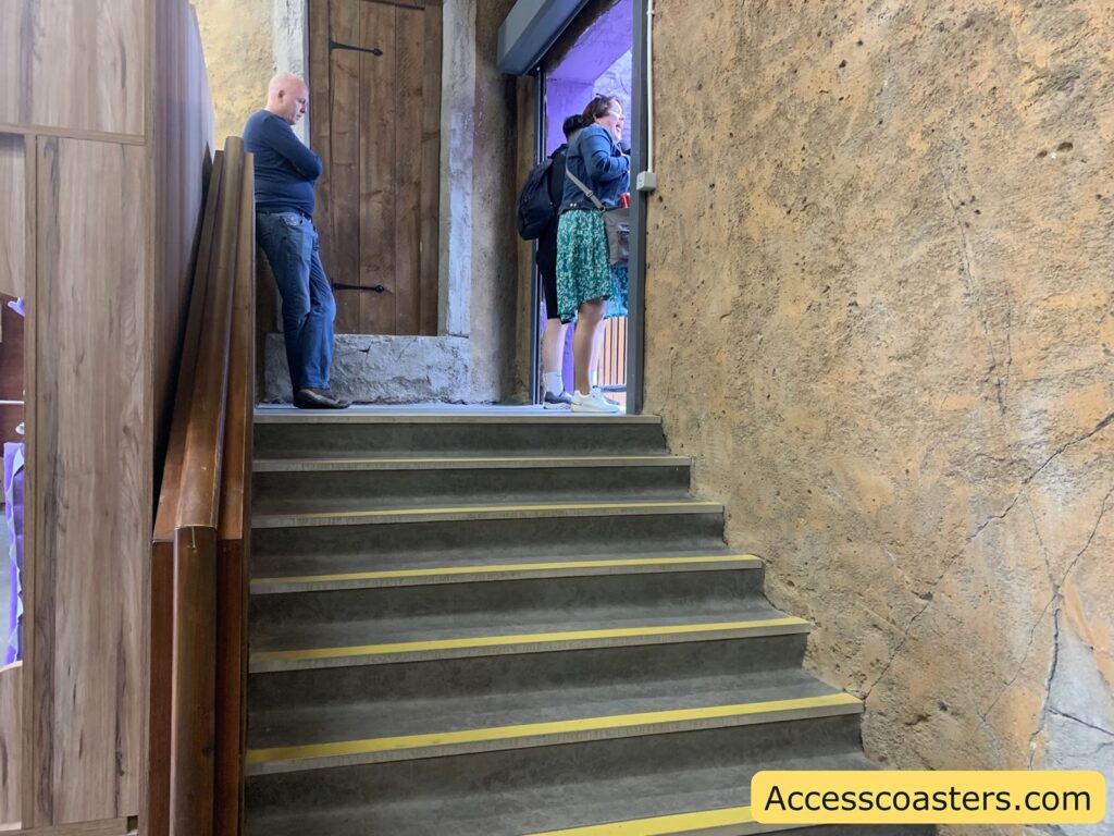 Concrete staircase with yellow safety strips on each step, leading up to a landing where two women are standing near a doorway and a man is leaning against the wall with his arms crossed. The walls are textured and rustic, with one side made of wood and the other of rough stone or plaster. There is a wooden handrail on the left side of the stairs