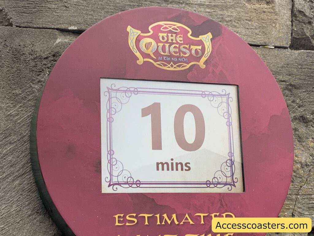 Theme park queue area with purple textured walls, black railings, and patterned tile flooring. Several people are standing in line, some facing forward and others looking at their phones. The lighting is dim with a blue glow in the background.