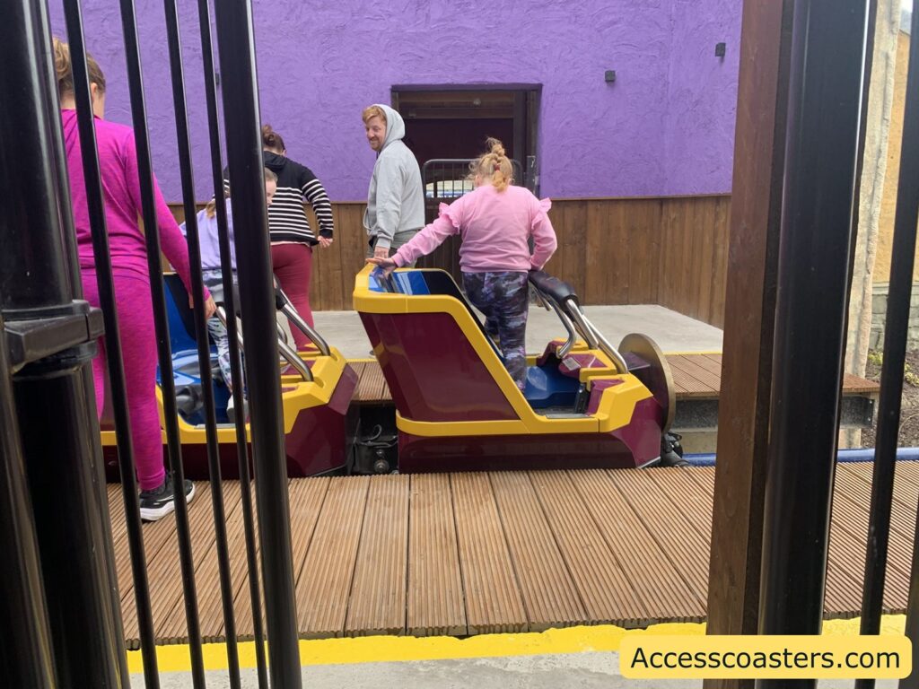 Amusement park ride loading area with people boarding. The image shows several people, mostly dressed in casual clothing like hoodies and leggings, getting into brightly colored ride vehicles. The vehicles are maroon and yellow with blue seats and metal safety bars. The ground is wooden decking, and there are black metal railings in the foreground.