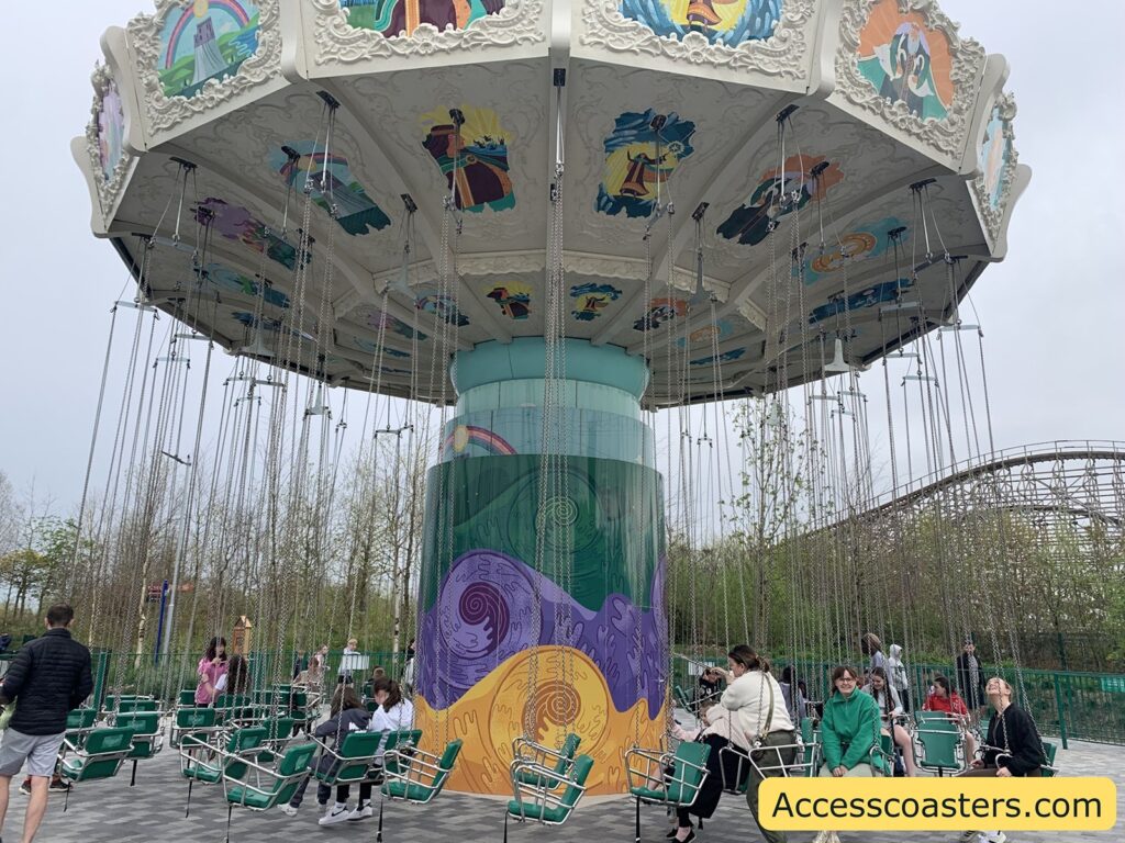 Amusement park swing ride with green seats hanging from chains, arranged in a circle around a central decorated column. The top canopy is adorned with colorful painted panels featuring various characters and scenes. Several people are seated and waiting for the ride to start, while others stand nearby. 