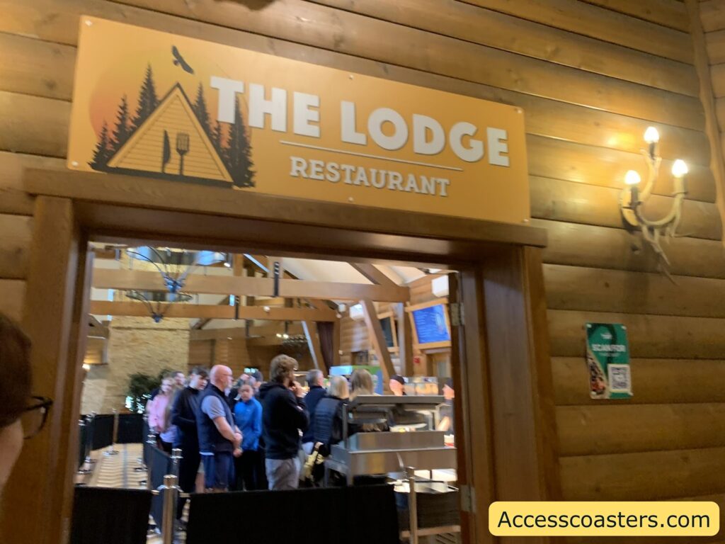 Entrance to a rustic-themed restaurant called "The Lodge Restaurant." The sign above the doorway is orange with white text and features a graphic of a cabin, pine trees, a fork, and a bird flying in front of a sunset. The walls are made of wooden logs, enhancing the lodge atmosphere. Inside, there is a line of people waiting