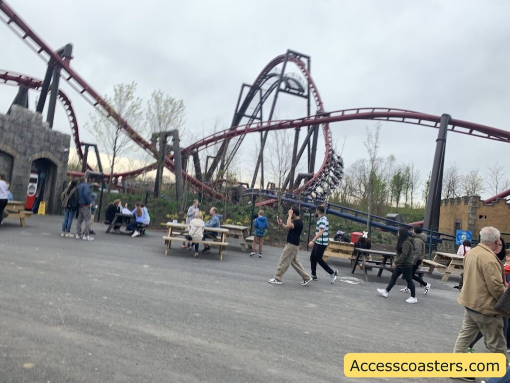 Amusement park scene with a roller coaster. The image shows a large, looping roller coaster with maroon-colored tracks and black supports in the background. The coaster train, filled with riders, is visible on the track, going through a turn. In the foreground, there are several wooden picnic tables where people are sitting and talking. 