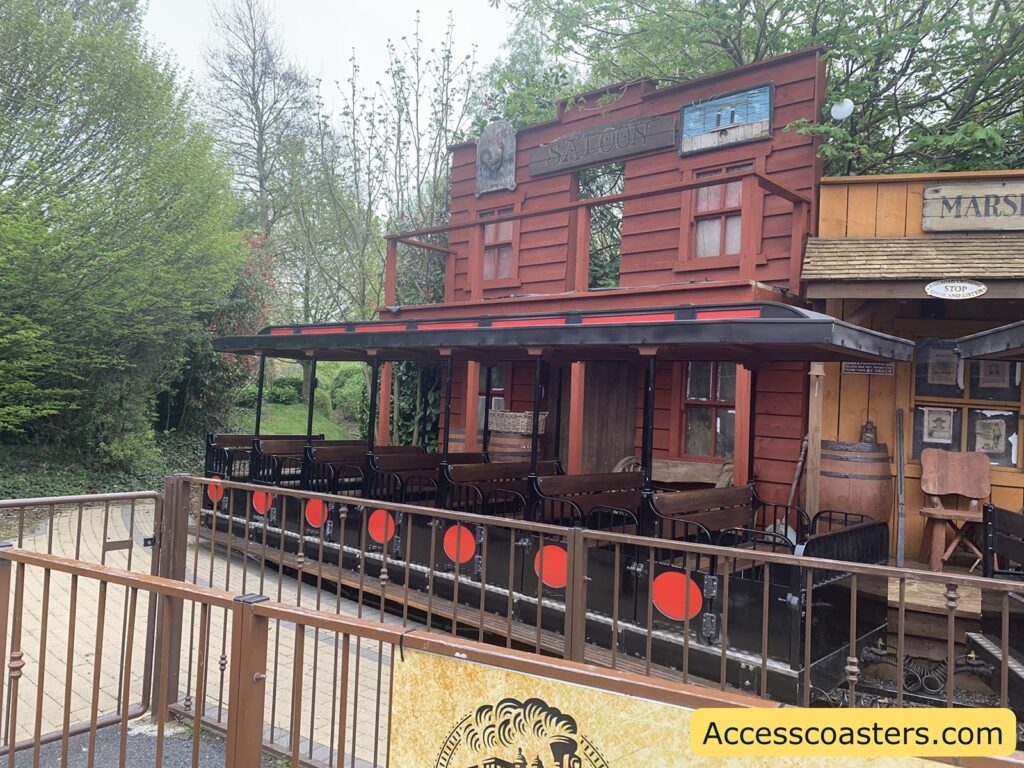 A western-themed amusement park ride station. The image shows a ride vehicle designed to look like an old-fashioned train carriage, parked in front of a wooden building facade styled as a saloon. The saloon has a rustic red exterior with a sign reading "SALOON" above the door and windows. Next to it, there is another building labeled "MARSHAL" with wanted posters and barrels outside, enhancing the Wild West atmosphere. The ride vehicle has bench seating and is surrounded by a safety fence with red circular markers.