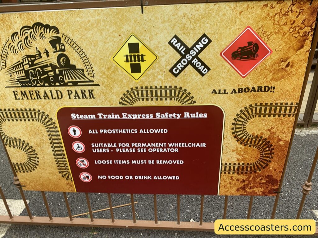Sign at a theme park entrance for a steam train ride. The background is a tan color with a stylized drawing of a steam locomotive and train tracks. The top left says "EMERALD PARK" next to the train illustration. There are three signs above the rules: a yellow diamond with a railroad track symbol, a black and white "RAIL CROSSING ROAD" sign in an X shape, and a red diamond with a black train icon. To the right, it says "ALL ABOARD!!" in bold letters.

Below, a maroon box with white text lists "Steam Train Express Safety Rules":
- ALL PROSTHETICS ALLOWED
- SUITABLE FOR PERMANENT WHEELCHAIR USERS - PLEASE SEE OPERATOR
- LOOSE ITEMS MUST BE REMOVED
- NO FOOD OR DRINK ALLOWED