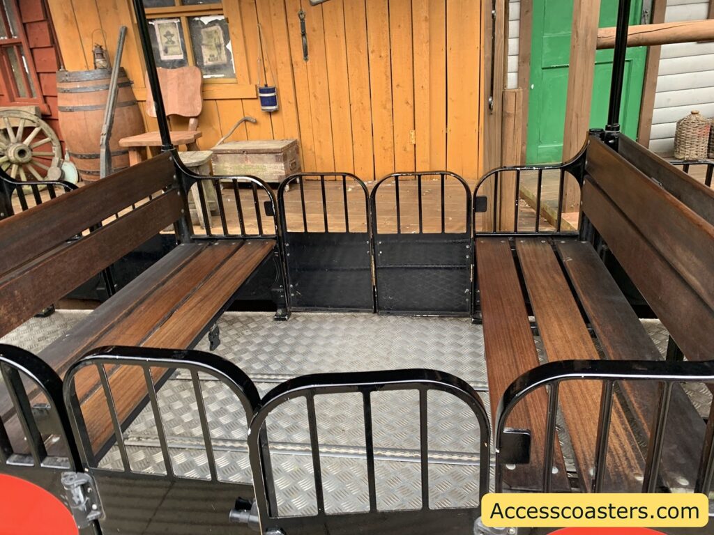 Wooden bench seating inside a theme park ride vehicle. The benches are arranged along the sides of the vehicle, facing inward toward each other, with black metal railings surrounding the seating area.  There is a track system on the floor to remove the seats for wheelchair access 