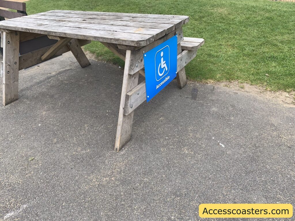 Wooden picnic table on a paved surface, with a blue sign attached to one side showing the white wheelchair accessibility symbol and the word "Accessible." The table is designed with an overhang on one end to allow wheelchair users to sit comfortably.