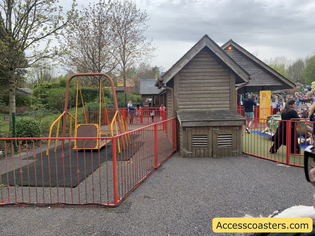 Playground scene with a fenced-in swing. The image shows a playground area with a large, accessible swing designed for children with disabilities. The swing is surrounded by a red metal fence and sits on a rubber safety mat. To the right, there is a wooden playhouse structure, and behind it, more people and children are visible, some playing and others standing around.