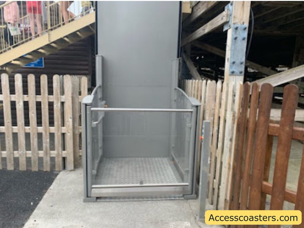A small outdoor wheelchair lift positioned between wooden fences. The lift has metal railings on three sides and a textured metal floor. There is a control panel on the right side of the entrance. The lift is located at the base of a staircase, which is visible in the background along with people standing on it.
