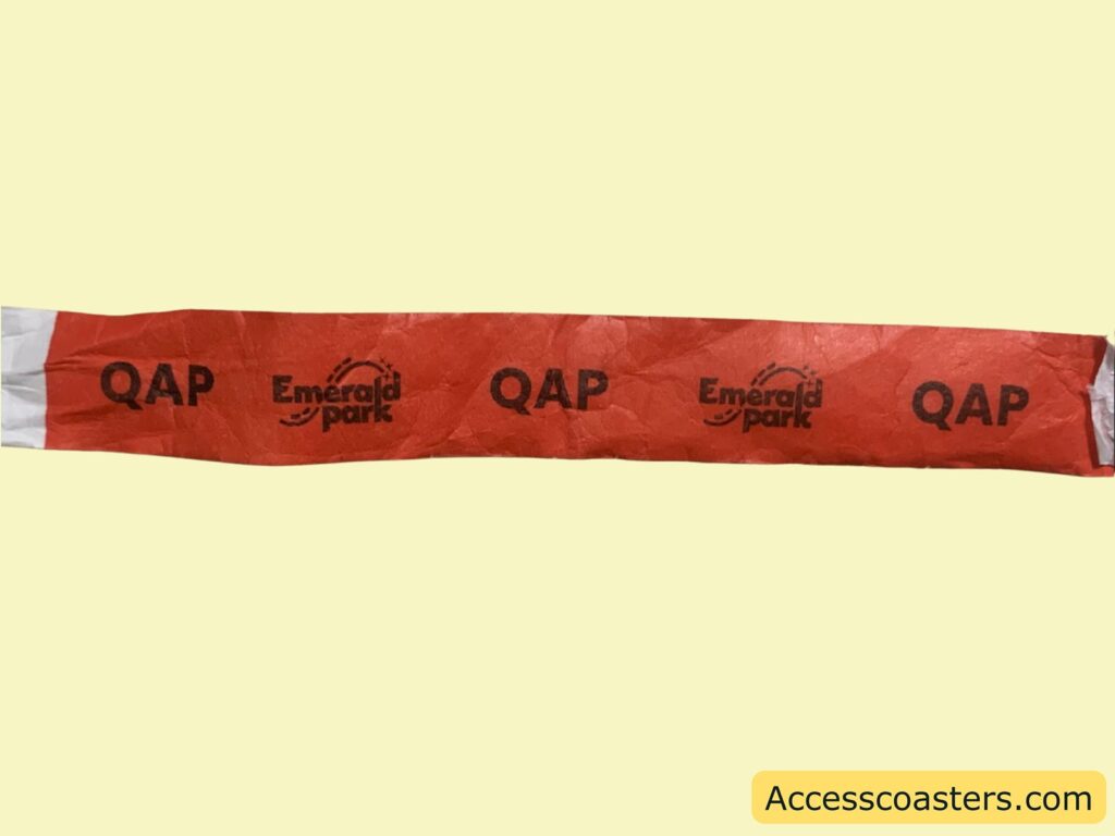 Red  paper wristband with black text. The wristband has the words "QAP" and "Emerald park" printed repeatedly along its length. The "Emerald park" text is accompanied by a small logo with stars. 