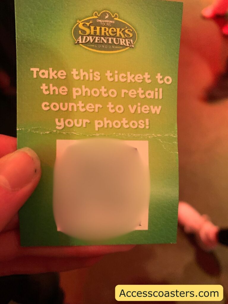 A green ticket being held between someone's fingers. The top of the ticket features the logo for "DreamWorks Tours Shrek's Adventure! London." Below the logo, large white text reads: "Take this ticket to the photo retail counter to view your photos!" In the center of the ticket, there is a rectangular area that is blurred out, likely concealing a barcode or unique code.