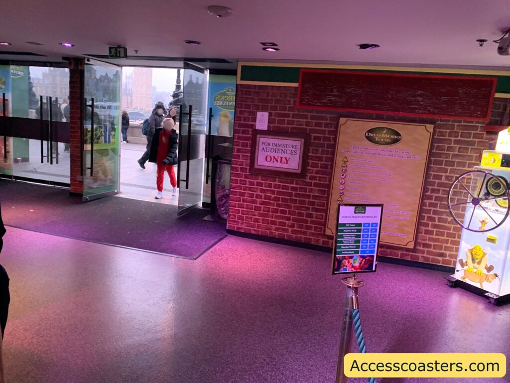 Indoor entrance area with glass doors open to the outside, letting in daylight. The interior has purple lighting on the floor, brick-patterned walls, and several signs. On the right, there is a large DreamWorks Tours sign with warnings and information.