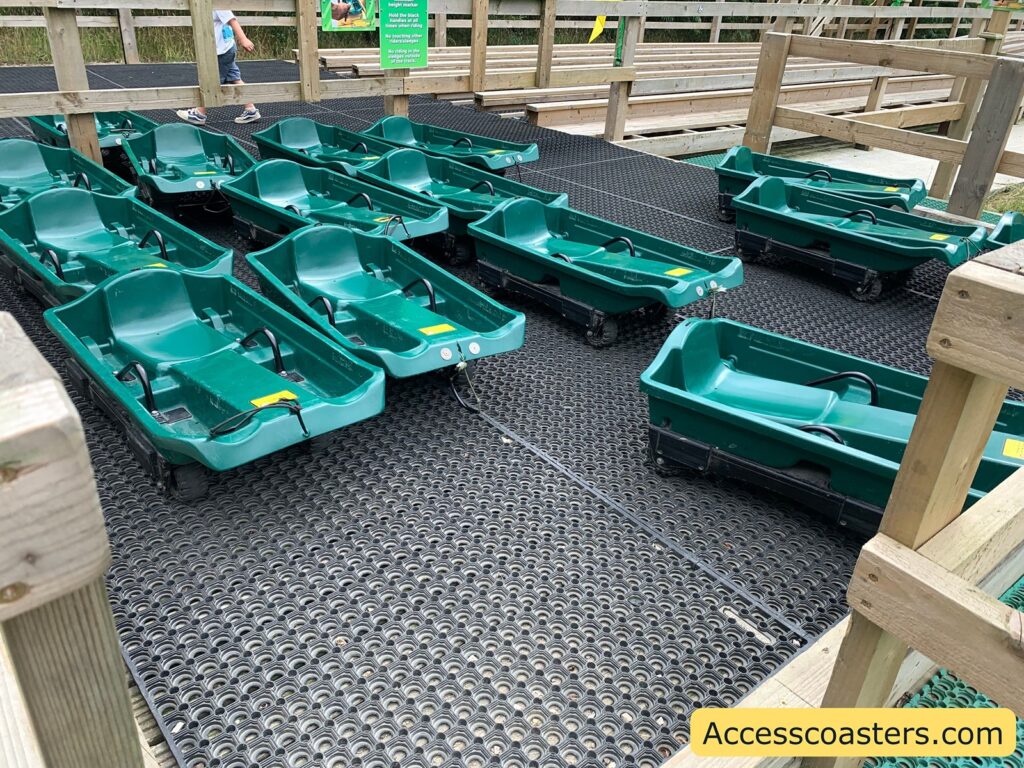 Green plastic sled-like carts lined up on a black rubber mat, surrounded by wooden railings. Each cart has a seat and handles,