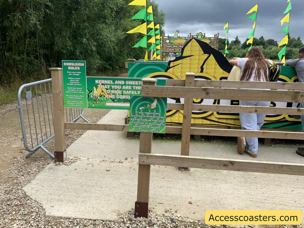 Outdoor amusement ride entrance area with wooden fencing, green and yellow signage, and colorful flags. The main sign features a large cartoon corn cob and the word "COBS" in big letters. 