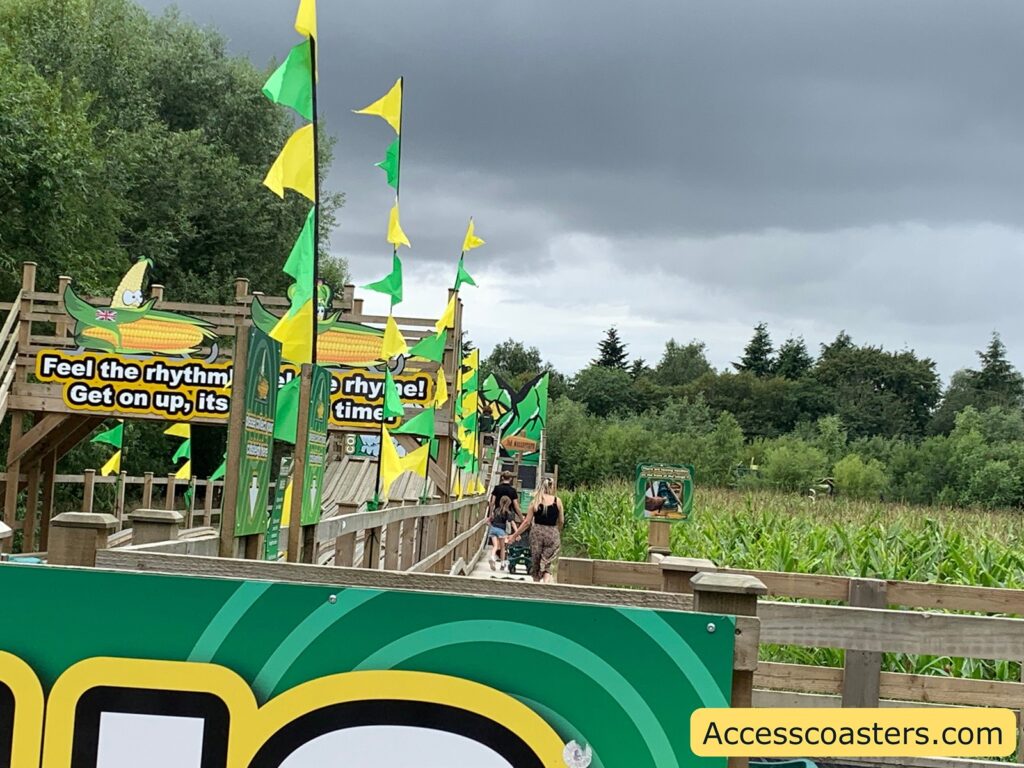 Outdoor walkway decorated with green and yellow flags, surrounded by lush greenery and cornfields. There are playful signs featuring cartoon corn cobs, one of which reads, "Feel the rhythm! Get on up, it's time!" Several people are walking along the wooden path