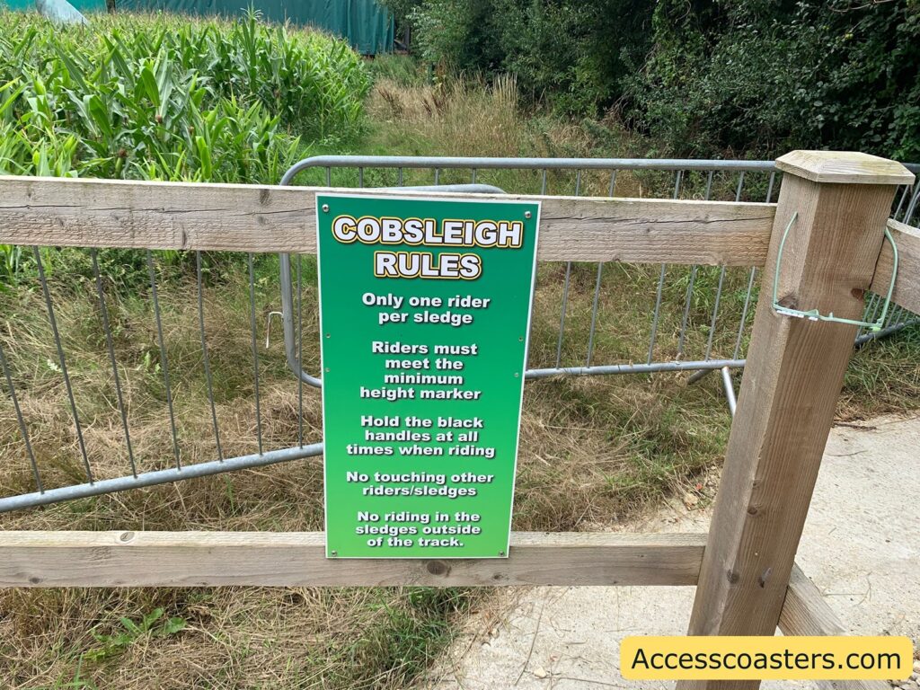 Green sign on a wooden fence with "COBSLEIGH RULES" in bold yellow and white letters at the top. The rules listed are:

Only one rider per sledge
Riders must meet the minimum height marker
Hold the black handles at all times when riding
No touching other riders/sledges
No riding in the sledges outside of the track