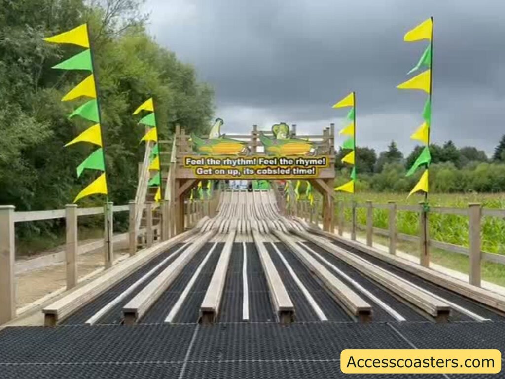 Outdoor multi-lane slide attraction with wooden lanes, surrounded by yellow and green flags on tall poles. At the top of the slide, there is a sign with a cartoon corn cob and the text: "Feel the rhythm! Feel the rhyme! Get on up, its cobsled time!" The slide is set in a rural area with trees and fields in the background under a cloudy sky.