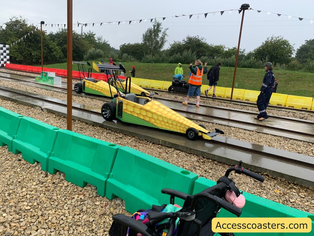 Outdoor racing track scene with two small, single-seat cars designed to look like ears of corn, complete with yellow kernels and green husks. The cars are positioned on parallel tracks, and several people, some in rain gear and high-visibility vests, are standing nearby. The ground is wet, indicating recent rain, and there are colorful plastic barriers (green, yellow, and red) along the track. In the foreground, there is a wheelchair with a pink cup attached. A checkered flag finish line is visible in the background,
