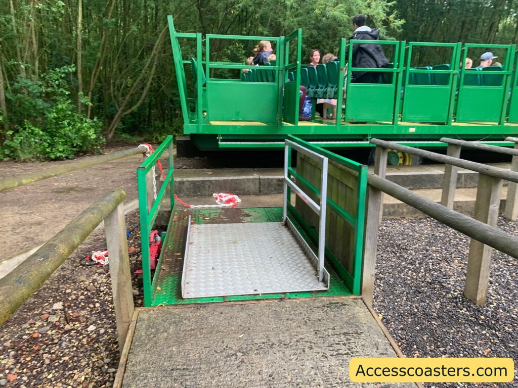 Accessible boarding ramp for a green amusement park ride. The ramp is made of metal with side railings and leads directly to the ride platform, which is at the same height as the ramp for easy access.