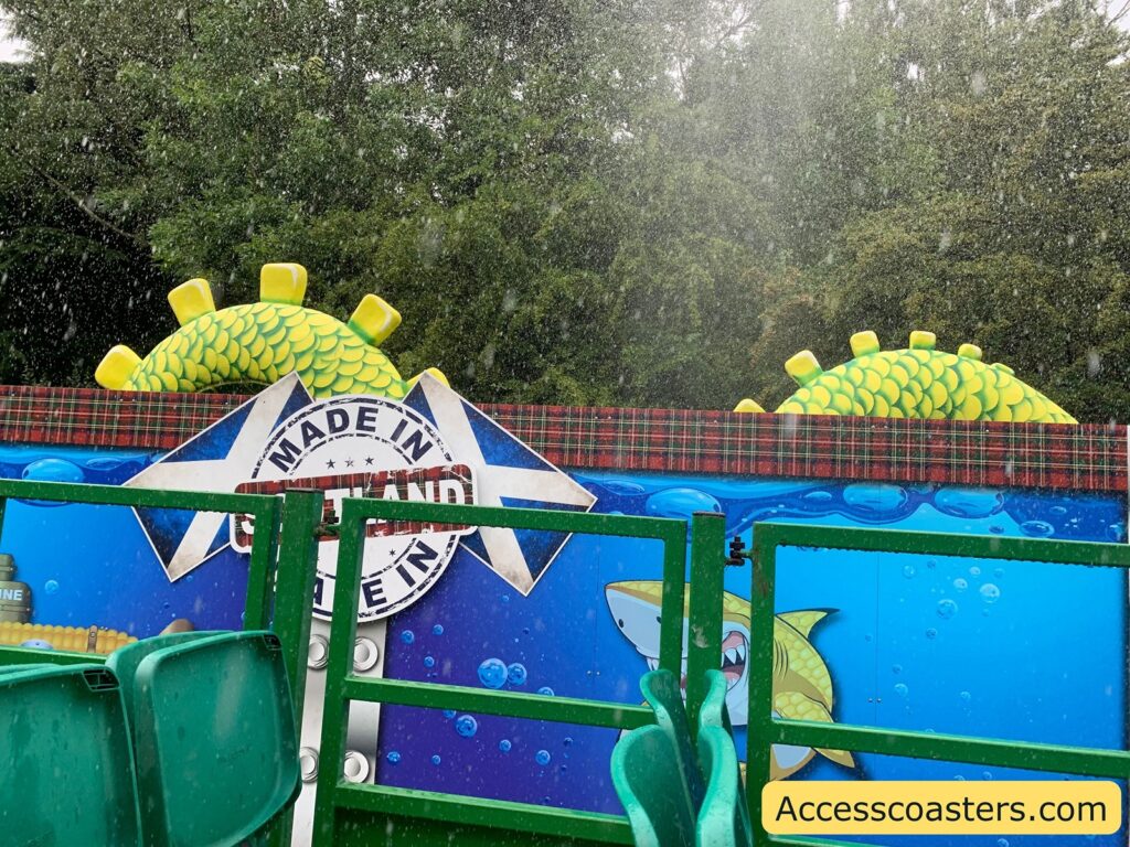 Outdoor amusement ride scene in the rain. The foreground features green plastic seats and a green metal safety rail. Behind the rail is a colorful mural with a blue underwater theme, including a cartoon yellow-green sea creature with scales and yellow spikes, and a smiling shark. The mural has a badge that reads "MADE IN" with "SCOTLAND" partially visible.
