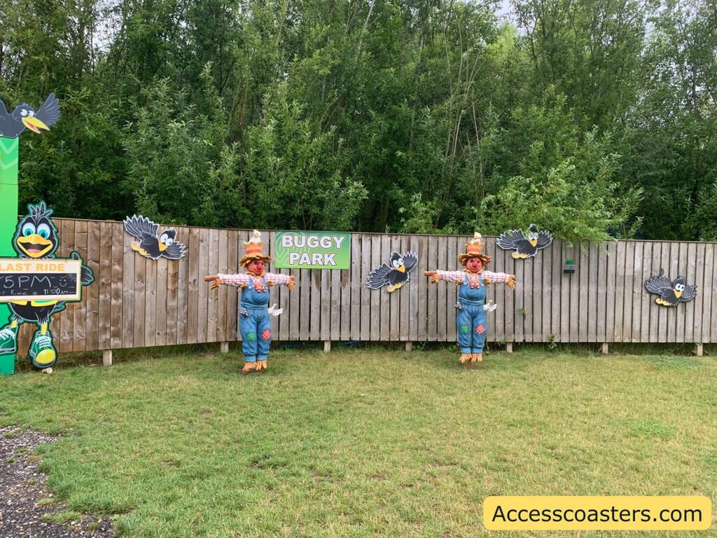 Outdoor scene at a children's amusement or theme park area. There is a wooden fence with cartoonish black birds attached to it, and two colorful scarecrow figures dressed in overalls, straw hats, and plaid shirts standing in front of the fence. A green sign on the fence reads "BUGGY PARK." 