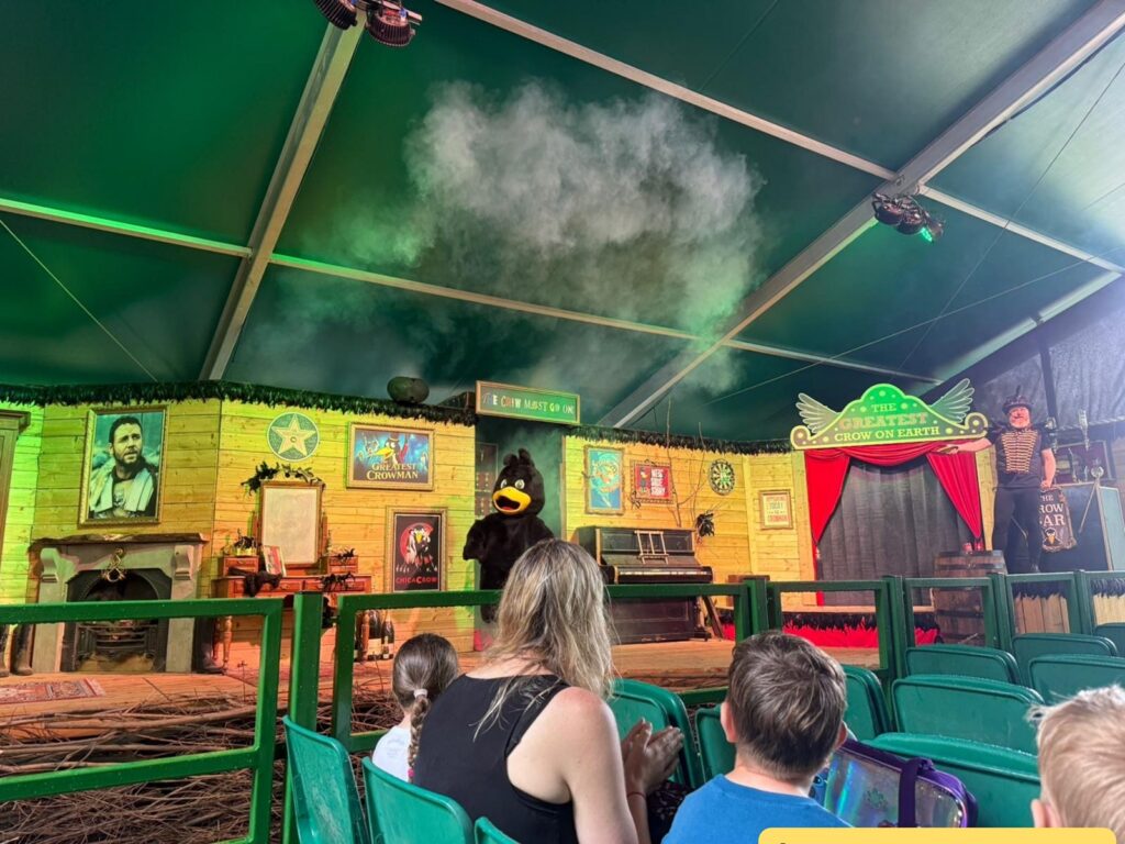 Indoor stage scene at a children's show. The stage is decorated like a cozy wooden room with various colorful posters, framed pictures, and props such as a fireplace, piano, and desk. There is a large green sign that reads "The Greatest Crow on Earth" above a small red-curtained area. On stage, a person in a black crow costume with a yellow beak stands near the center, while another performer dressed in a striped shirt and top hat stands to the right. Smoke or mist is coming from above the stage, 