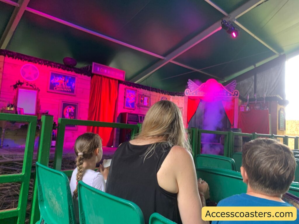 the tractor in the crow man house. The stage is decorated with red curtains, colorful posters, and a wooden backdrop, with purple and pink lighting creating a lively atmosphere. There is a section of the stage emitting purple smoke or mist, adding a sense of anticipation or theatrical effect.
