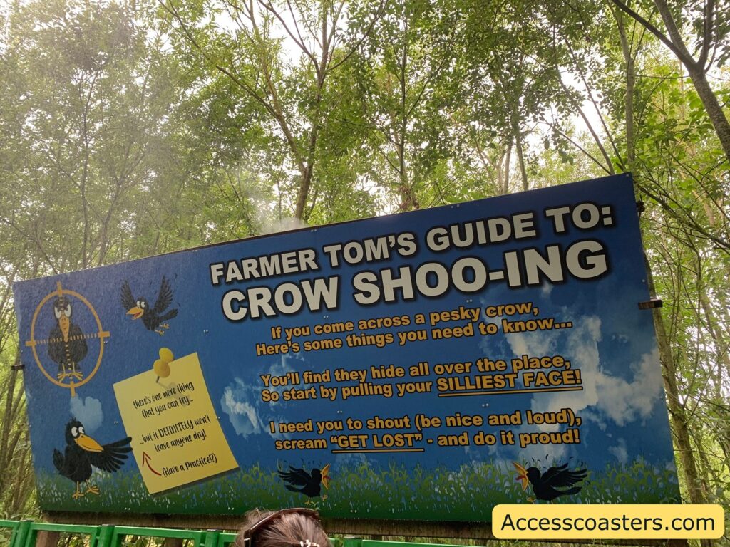 Large blue sign in a wooded area with the title "FARMER TOM'S GUIDE TO: CROW SHOO-ING." The sign gives playful instructions for scaring away crows, suggesting to pull your silliest face and shout "GET LOST" loudly and proudly. There are cartoon crows illustrated on the sign, one inside a target, and another holding a note that says, "there's one more thing that you can try - but it DEFINITELY won't leave anyone dry! (Have a Practice!)"
