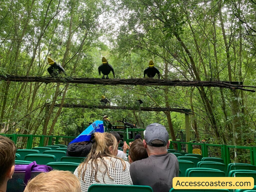 Outdoor amusement park ride scene with green seats and several people sitting, seen from behind. The ride is moving through a lush, wooded area with many trees. Overhead, there are large, artificial black birds with yellow beaks perched on wooden structures above the ride path. A staff member in a green uniform is visible ahead, standing near the front of the ride.