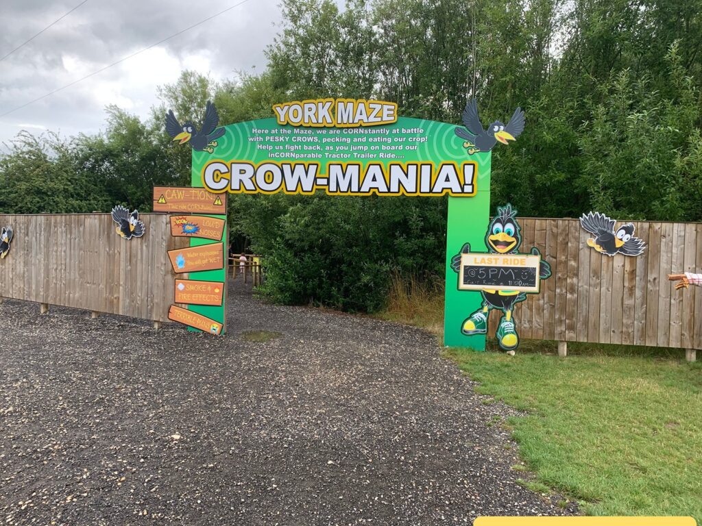 Entrance to an outdoor attraction called "York Maze CROW-MANIA!" The entrance is marked by a large, colorful sign with cartoon crows and a green background. There is a wooden fence on both sides, decorated with more cartoon crows. A sign on the left warns visitors about loud noises, water splashes, smoke and fire effects, and terrible puns. On the right, a cartoon crow holds a chalkboard sign that says "LAST RIDE @ 5PM! First Ride @ 11:00AM." 