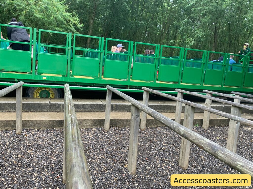 Outdoor queue area with wooden railings leading up to a green open-sided tractor ride. The tram is parked and has several people seated inside