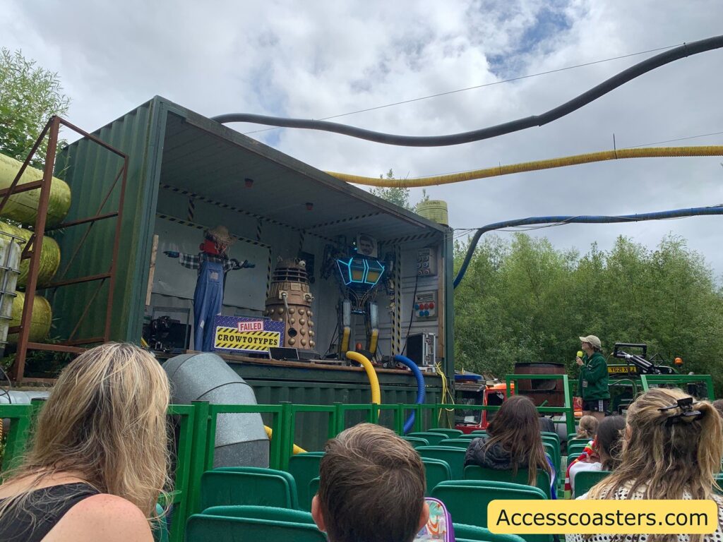 Outdoor stage show scene. The image shows an audience seated in green chairs, watching a performance set up inside an open shipping container. On the stage, there is a scarecrow figure dressed in overalls, a robot that resembles a Dalek from Doctor Who, and a person in a glowing, futuristic costume. There are large tubes and pipes connected to the container, giving a science experiment or mad scientist vibe. A sign on the stage reads "FAILED CROWOTYPES." To the right, a person with a microphone appears to be hosting or narrating the show.