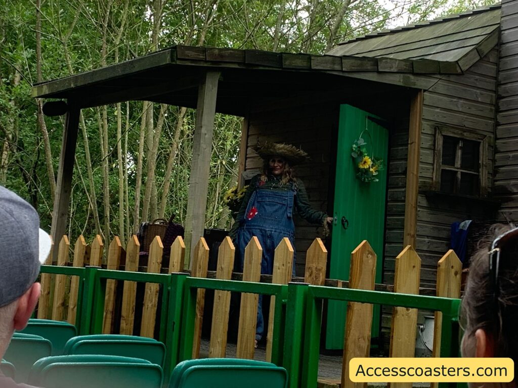 Outdoor stage scene with a rustic wooden house, a small porch, and a green door decorated with flowers. A person dressed as a scarecrow, wearing a straw hat and blue overalls, stands in the doorway. 