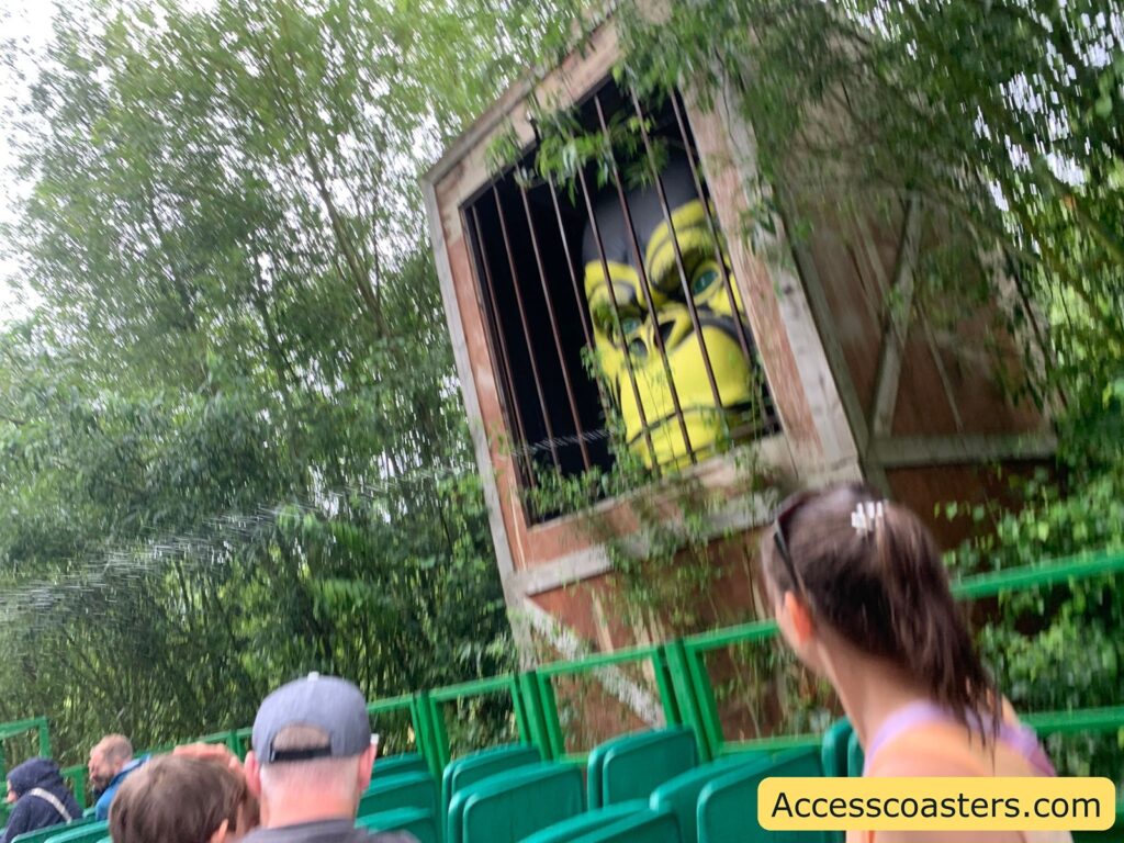 Outdoor amusement park or zoo setting with green seating and several people sitting and looking toward a large, cartoonish gorilla face behind bars in a wooden enclosure. The gorilla face is yellow and black, appearing as part of a themed attraction