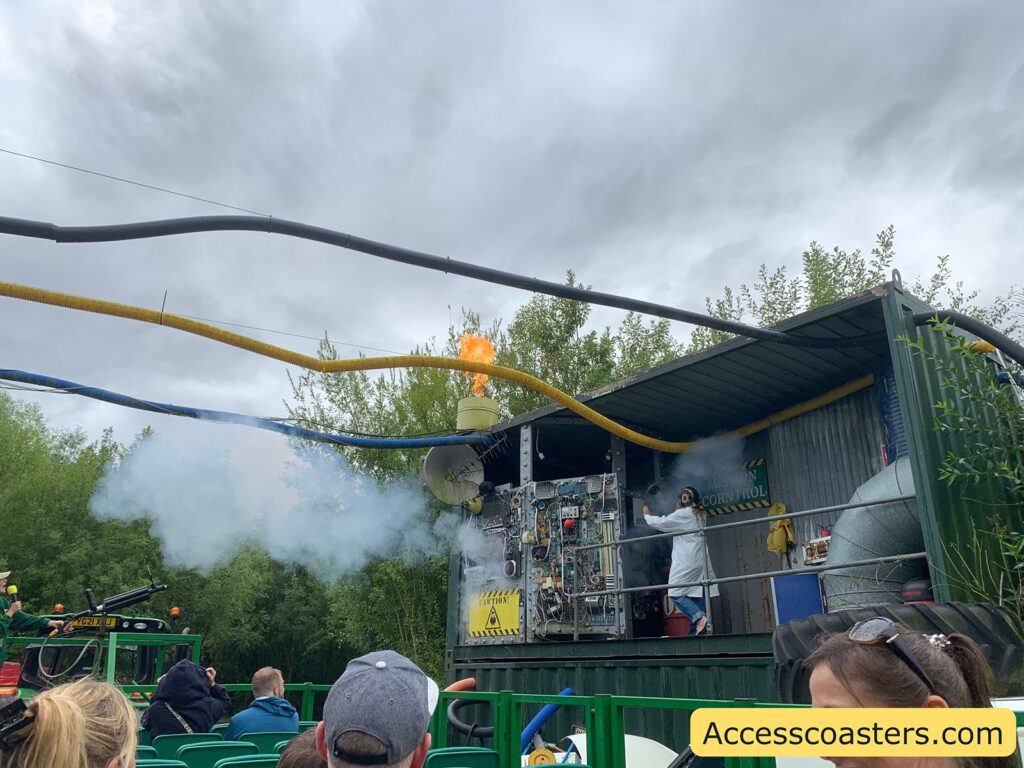 Outdoor theme park attraction scene. In the foreground, people are seated, facing a stage-like setup. The stage is designed to look like a quirky laboratory or control station, with various pipes, wires, and machinery. A person in a white lab coat and goggles is performing, possibly acting as a scientist. There is smoke coming from the setup and a burst of flame shooting upward