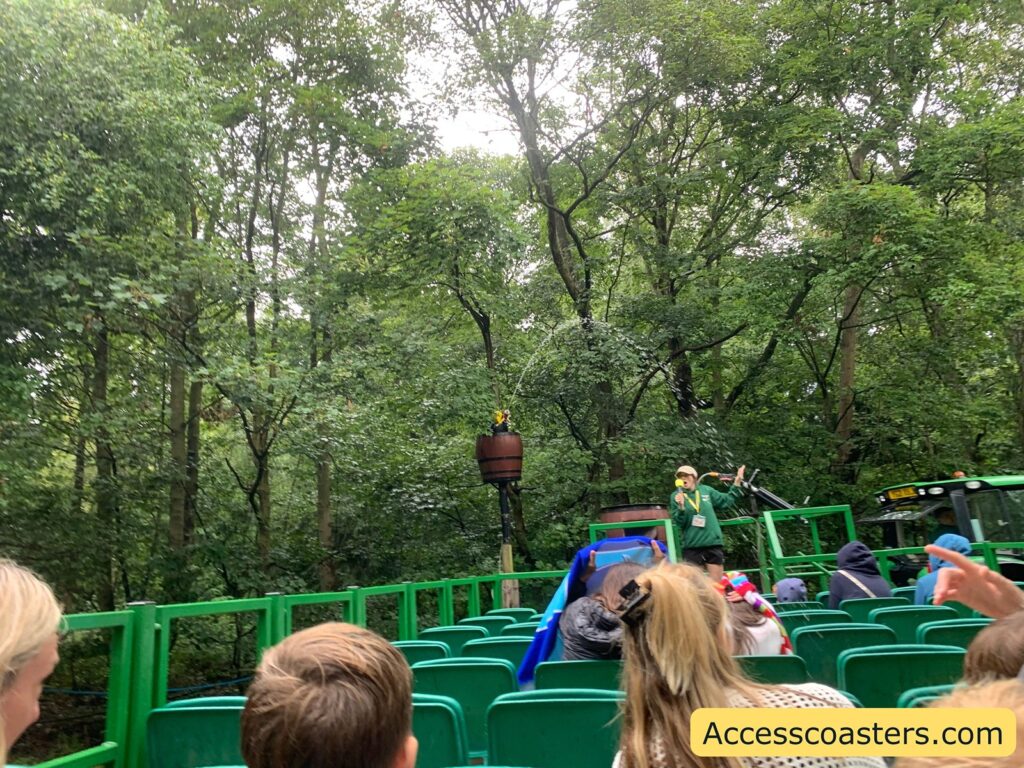 Outdoor ride scene in a forested area with green trees and foliage surrounding a group of people seated in green open-air tram or ride vehicle seats. A staff member in a green uniform and yellow cap stands at the front, speaking into a microphone. Water is spraying from a barrel elevated on a pole, creating an arc over the ride vehicle. 