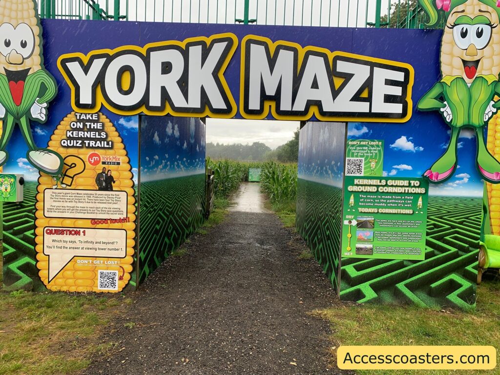 Entrance to a corn maze called "York Maze." The entrance is flanked by two cartoon corn characters, one on each side, both smiling and wearing green husks. The large sign above the entrance reads "YORK MAZE" in bold white letters with a yellow outline.

On the left side, there is a sign inviting visitors to take on the "Kernels Quiz Trail!" with a quiz question: "Which toy says, 'To infinity and beyond'? You'll find the answer at viewing tower number 1." There is also a QR code and a reminder not to get lost.

On the right side, there is a "Kernels Guide to Ground Conditions" explaining that the maze is made from a field of corn and can become muddy when wet. There is a visual indicator for today's ground conditions, and another QR code for help.

The path leads straight into the corn maze, with tall green corn plants visible ahead.