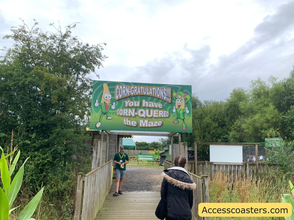 Outdoor scene at the exit of a corn maze. A large green sign above the wooden walkway reads: "CORN-GRATULATIONS!!! You have CORN-QUERED the Maze" with playful cartoon corn characters on either side