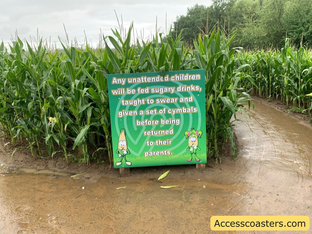 A humorous warning sign in a cornfield maze. The sign reads: "Any unattended children will be fed sugary drinks, taught to swear and given a set of cymbals before being returned to their parents." The sign has a green background with cartoon corn characters on the bottom corners.