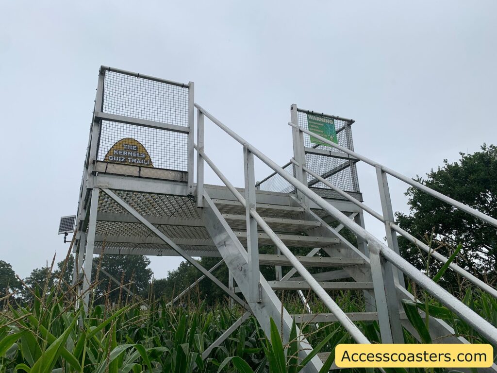 Metal observation platform with stairs, surrounded by tall green corn plants. At the top of the platform, there are two signs: one on the left reads "THE KERNELS QUIZ TRAIL" in yellow, and the one on the right is green and says "WARNING" at the top,