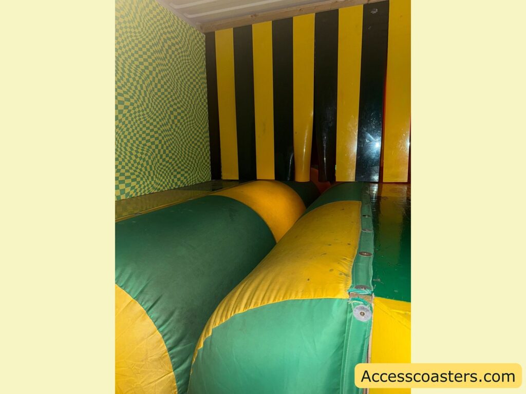 nflatable obstacle course section. The image shows large, padded, green and yellow inflatable barriers running horizontally, with a wall at the end featuring alternating vertical black and yellow stripes. 